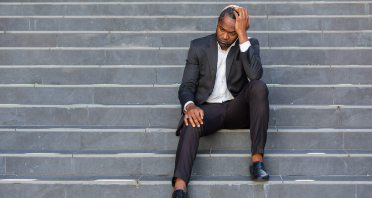 Sad african american businessman sitting on stairs outside office building, man in business suit