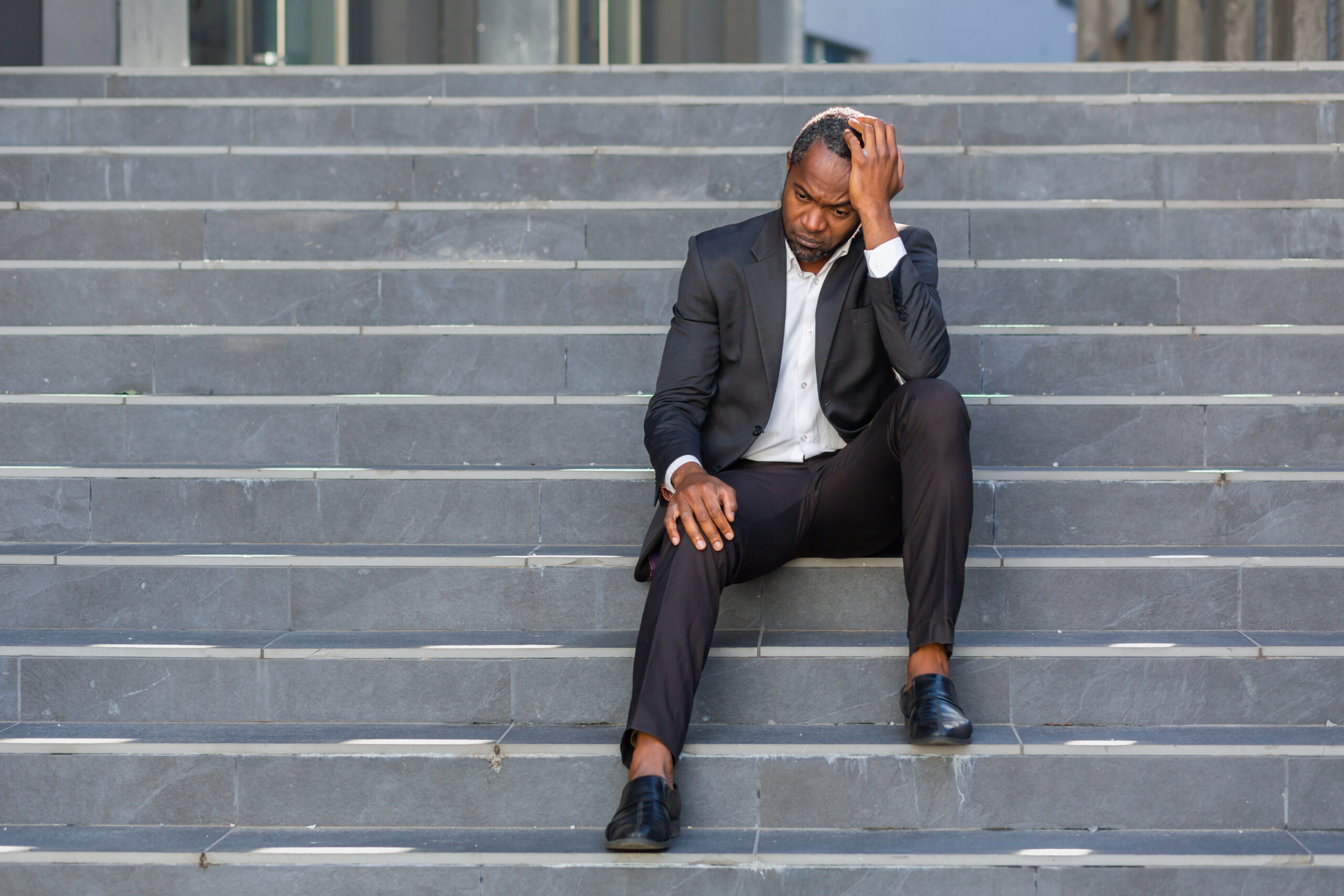 Sad african american businessman sitting on stairs outside office building, man in business suit